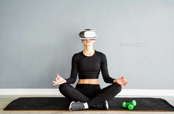 Young blonde woman in sport clothes wearing virtual reality glasses meditating on fitness mat