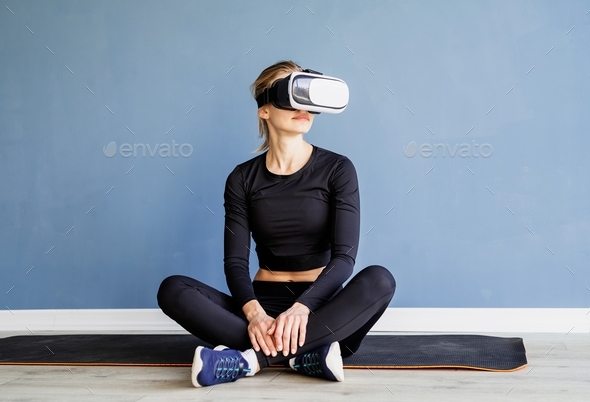 Young blonde woman in sport clothes wearing virtual reality glasses sitting at fitness mat using VR