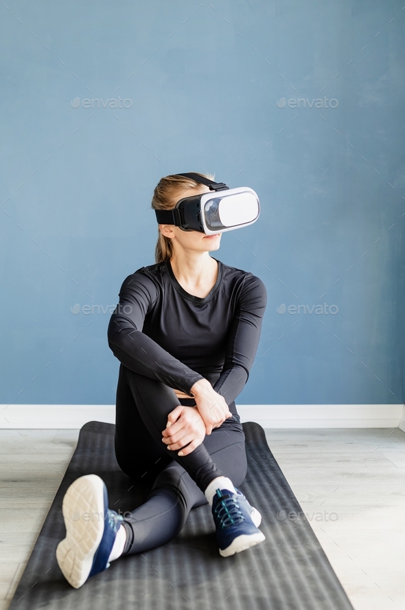Young blonde woman in sport clothes wearing virtual reality glasses sitting on fitness mat