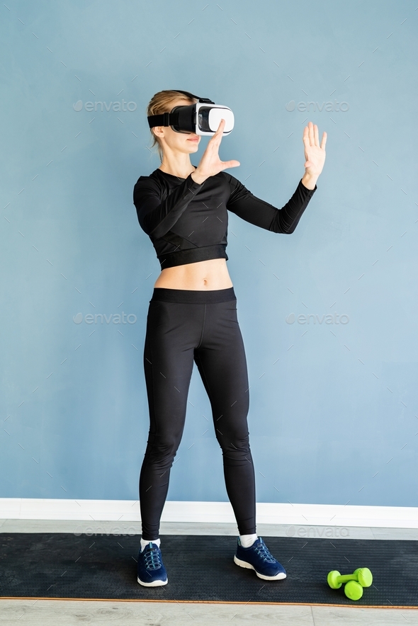 Young blonde woman in sport clothes wearing virtual reality glasses standing at fitness mat using VR