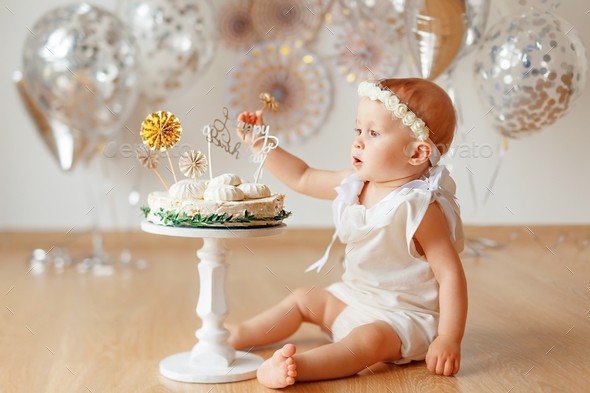 Little baby girl in white dress sitting on floor near birthday cake
