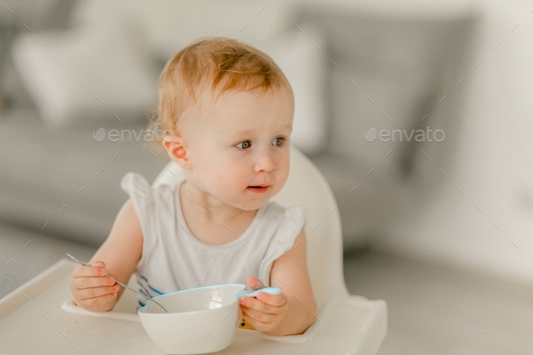 little girl under age of 1 year in bright kitchen in white highchair sits and eats from white plate