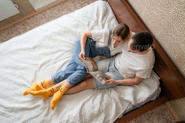 young couple lie on bed read magazine, spend time together