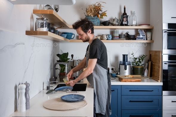 A man does housework. A young man washes dishes in the kitchen in a Scandinavian design