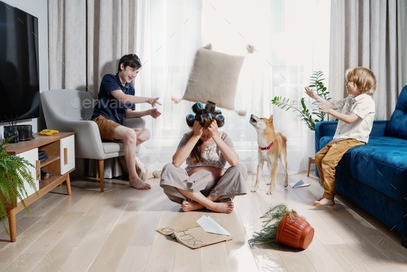 Two boys engage in pillow fight while their mother trying to keep calm in middle of family chaos