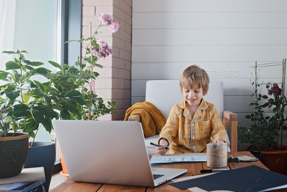 A boy in yellow pajamas learns to draw, cut and glue during an online lesson on the loggia