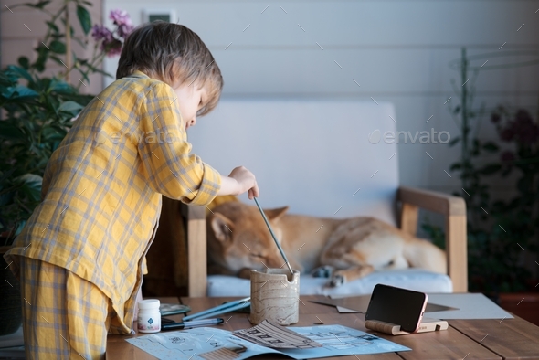 A boy in yellow pajamas learns to draw, cut and glue during an online lesson on the loggia