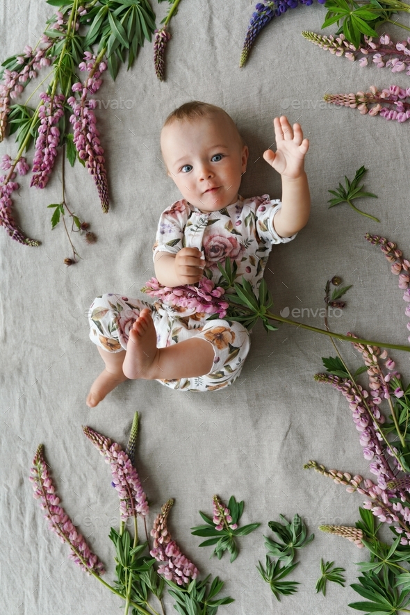 The cutest baby in calico clothes with floral print lying on linen textile blanket with pink lupins