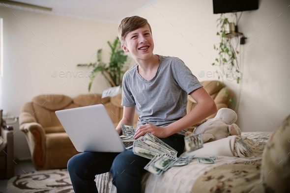 Laughing teenager with a laptop in a pile of banknotes. Earnings of schoolchildren online