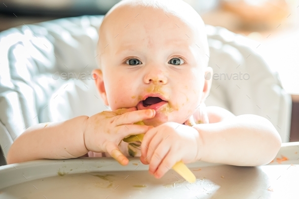 Messy Baby holds spoon with two hands sitting in kitchen at table after dinner. Complementary food