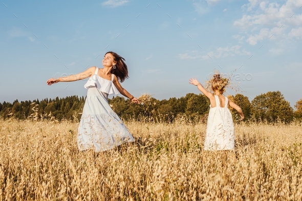 Beautiful young mother and her daughter dance in field. White clothes, freedom concept. Front view.