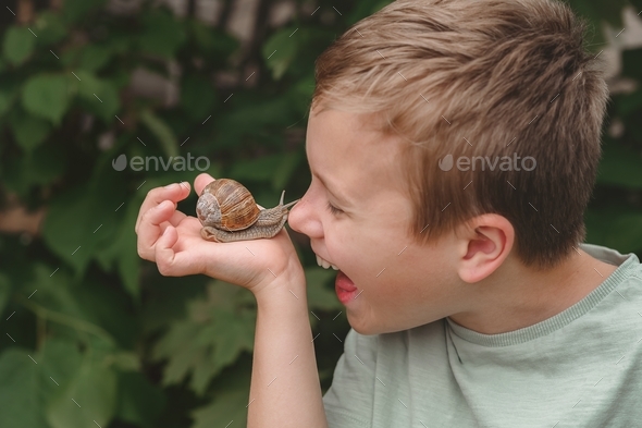 A happy child holds a big garden snail in hand and touches it with his nose in the park. Close up