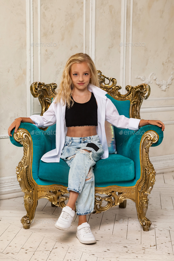 Little girl 6-7 years old blonde princess model in stylish clothe sitting on green armchair