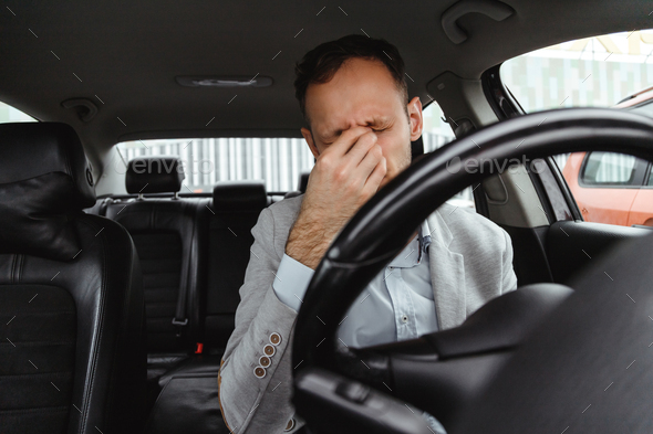 Tired man driver in suit rubbing his eyes behind steering wheel of car