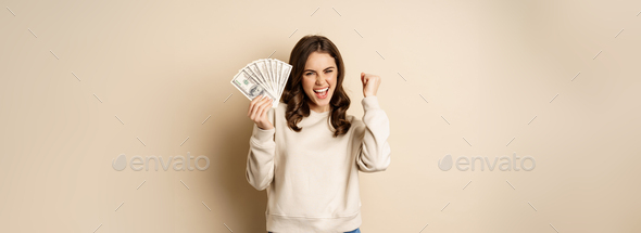 Enthusiastic modern woman winning money, got cash, celebrating and shouting of joy, standing