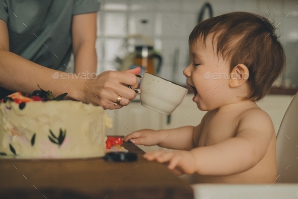 a woman\'s hand with a wedding ring waters a baby sitting in a chair at the table from a mug