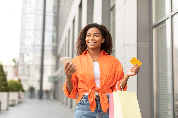 Glad happy millennial black lady in casual with smartphone, credit card and a lot of packages