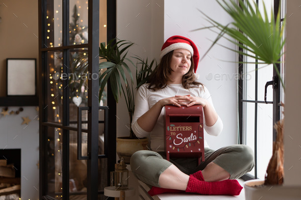 Dreaming smiling woman in red hat holding posrt box of letters to santa at home