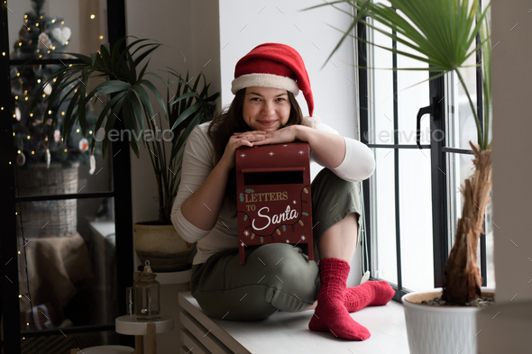 Funny caucasian woman in red hat with posrt box of letters to santa at home