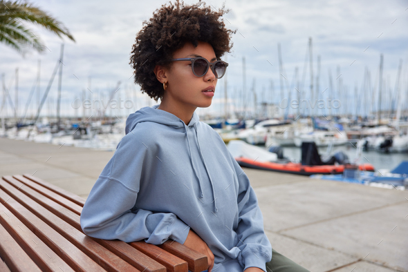 Horizontal shot of pensive Afro American woman dressed in hoodie and sunglasses sits on wooden bench