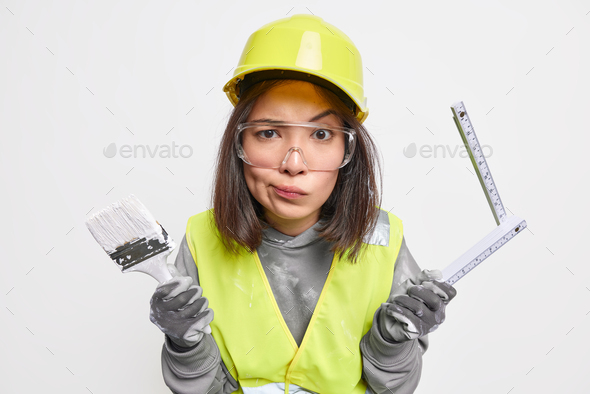Discontent brunette Asian woman maintenance worker holds painting brush and tape measure smirks face