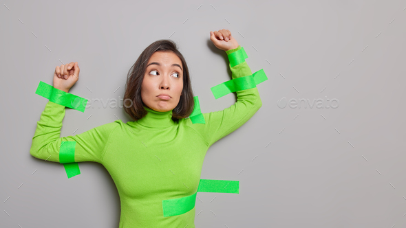 Frustrated unhappy Asian woman in green turtleneck plasted to grey wall keeps arms raised has discon