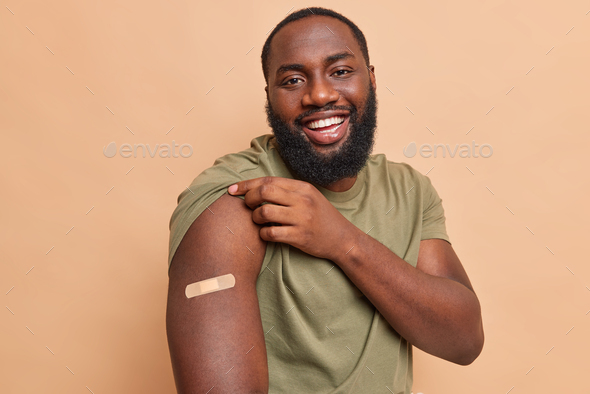 Cheerful dark skinned man shows adhesive plaster on shoulder after getting coronavirus vaccine feels