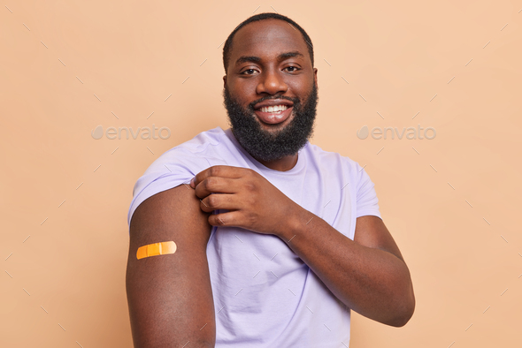 Happy dark skinned bearded man shows arm with adhesive plaster got coronavirus vaccine feels safe sm