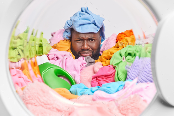 Domestic work and housekeeping concept. Unhappy dark skinned man covered with multicolored laundry l