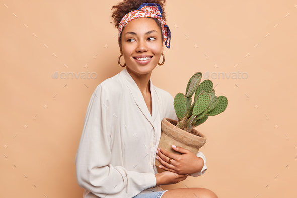 Pretty dark skinned curly haired woman sits against beige background looks happily away holds pot ot