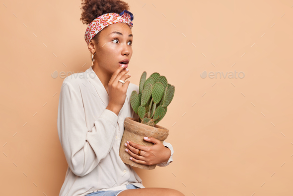 Sideways shot of surprised African American woman stares impressed holds breath holds potted cactus