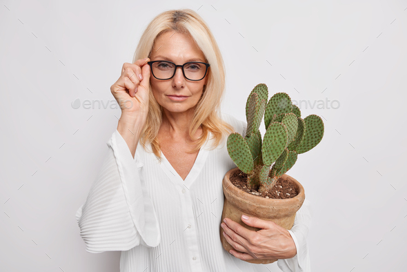 Serious middle aged woman with blonde hair keeps hand on rim of spectacles looks attentively at came