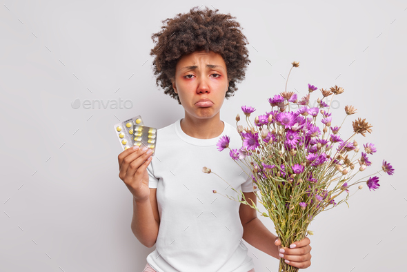 Puzzled unhappy African American woman holds bouquet of wildflowers and pills against allergy has re