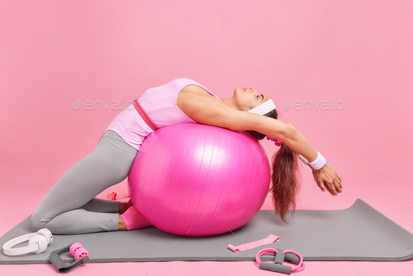 Horizontal shot of active slim woman leans over fitness ball does gymnastic at home dressed in bodys