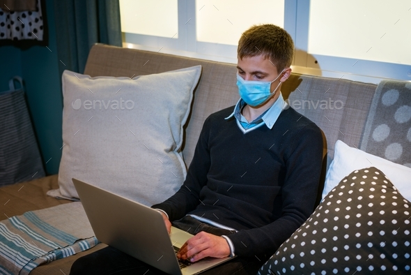 young caucasian man sitting at home on sofa with laptop in medical mask