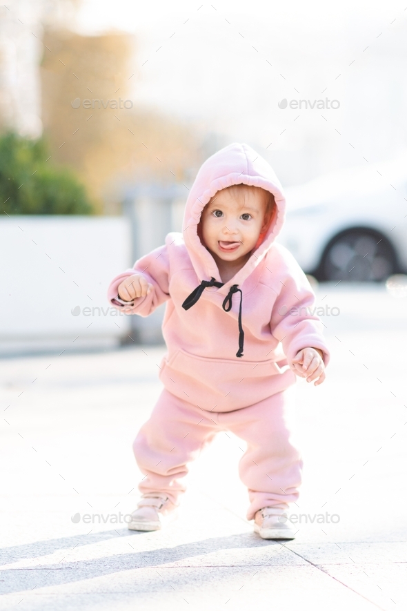 Cute baby girl wear trendy pink sport suit and hoody over city street outdoors. Childhood