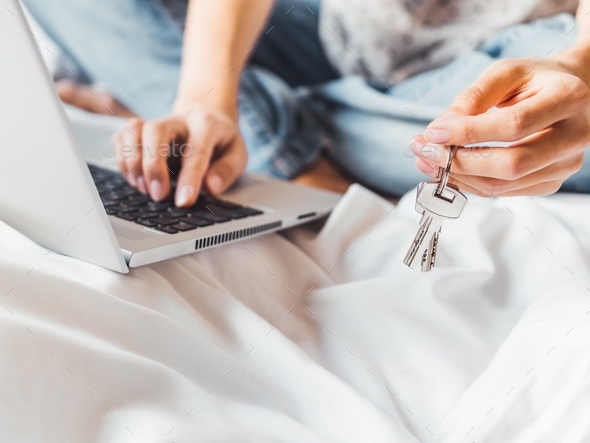 Woman uses laptop in bed to pay bills or book a hotel room. Metal door keys in her hand.
