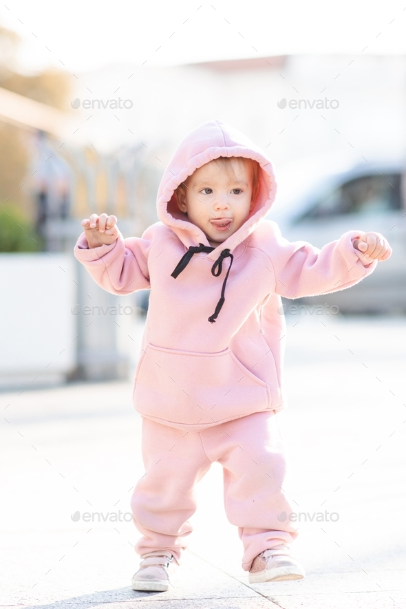 Cute baby girl wear trendy pink sport suit and hoody over city street outdoors. Childhood