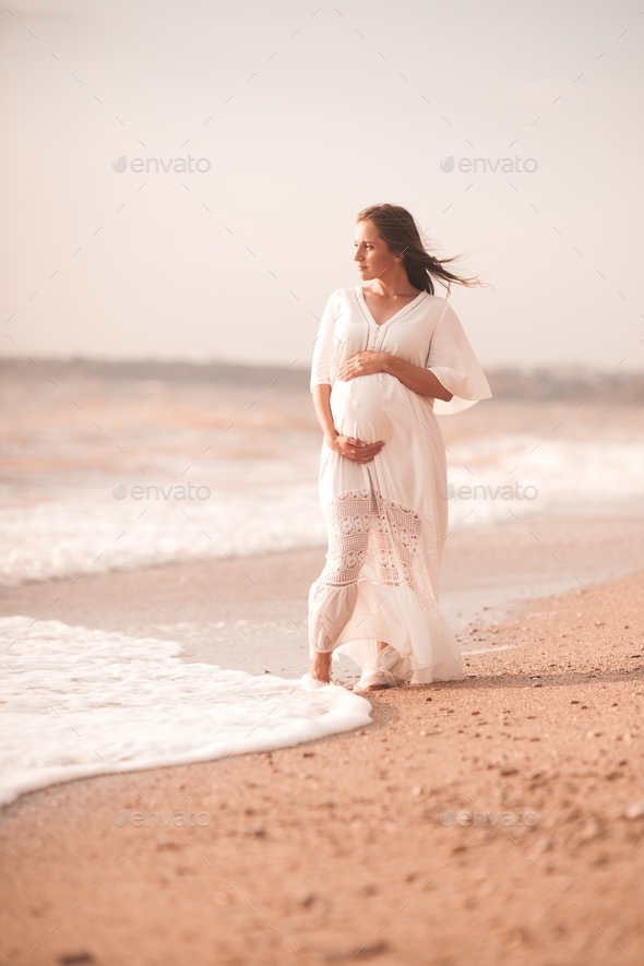 Blonde pregnant woman 24-26 year old wearing white dress holding tummy walking at beach outdoors.