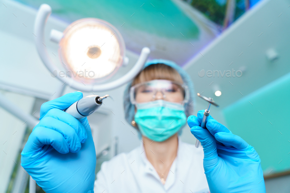 Dentist in a surgical mask, holding an angle mirror and a drill, ready to work.