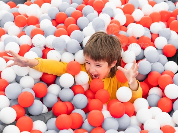 Laughing boy plays in ball pit. White and red plastic balls in dry paddling pool. Leisure activity.