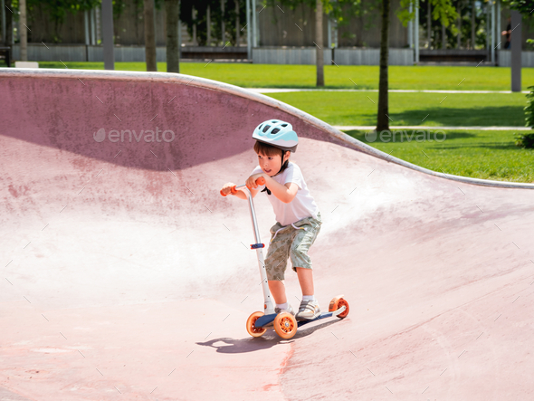 Boy trains to ride a kick scooter in concrete skate park. Active recreation at summer vacations.