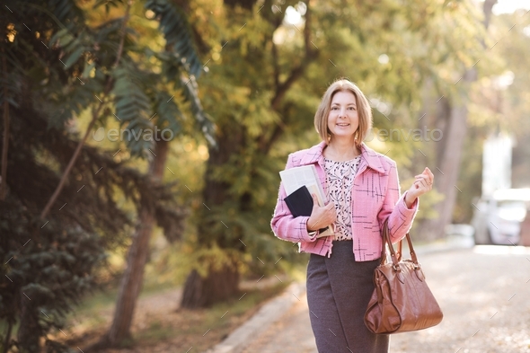 Stylish business mature woman 50-55 year old holding notebook and bag wearing suit walking in city