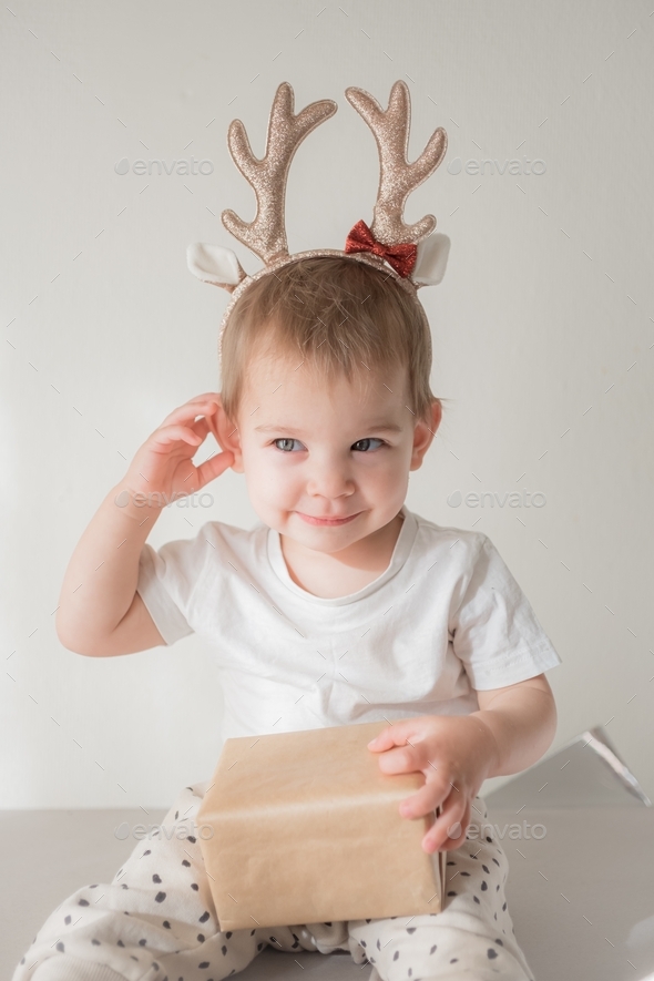 Little toddler girl in gender neutral clothes in rain deer costume horns new year with present