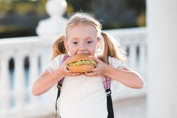 Kid girl 5-6 year old eating burger outdoors. Focus on sandwich. Good morning. Breakfast time.