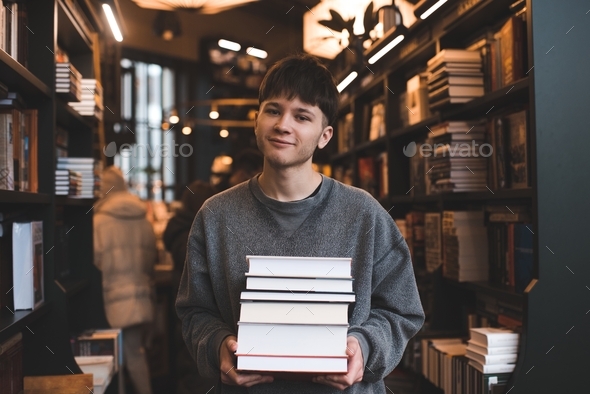 Happy handsome teenage guy holding paper books standing in shop over bookshelf. Young man student