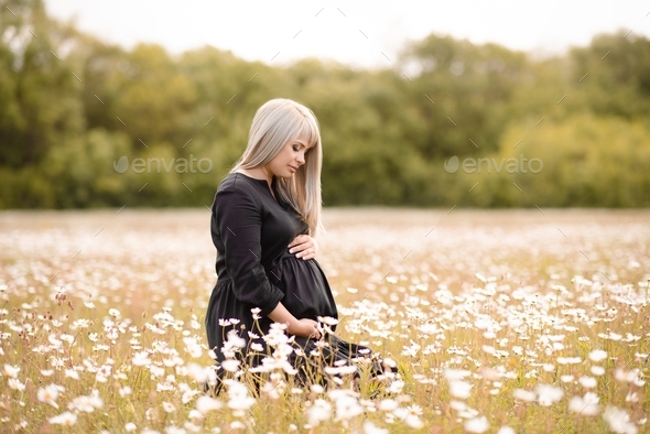 Cute pregnant woman holding belly sit in daisy meadow over nature background with flowers