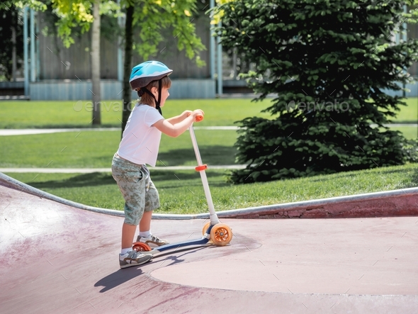 Boy trains to ride a kick scooter in concrete skate park. Active recreation at summer vacations.