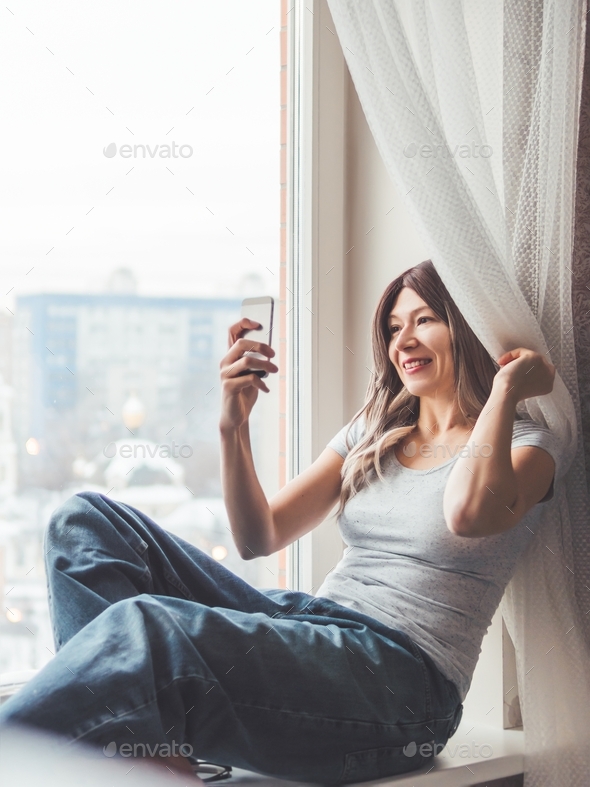 Woman with curly hair poses for selfie on windowsill. Pretty woman makes  self-portrait on smartphone Stock Photo by aksenovko