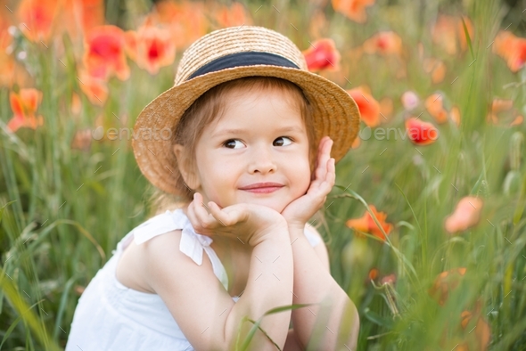 Cute funny little child girl wear hat sit in poppy meadow with red poppies outdoor. Springtime.